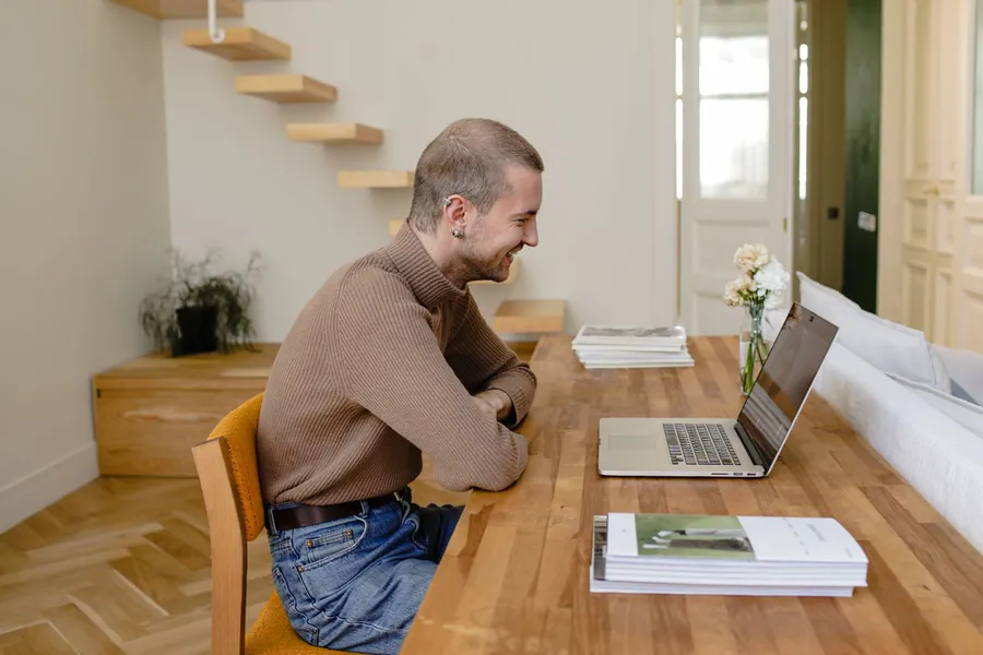 A man at a kitchen table, smiling at his laptop