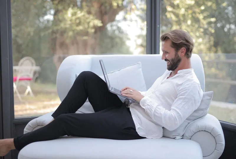 A man relaxing on a sofa by a window, using a laptop